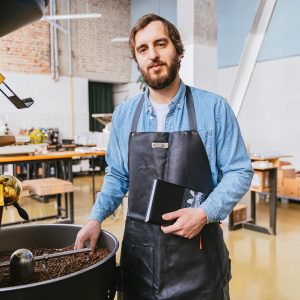 A bearded master roaster stands by a coffee roasting machine, holding a notebook in an industrial workshop.