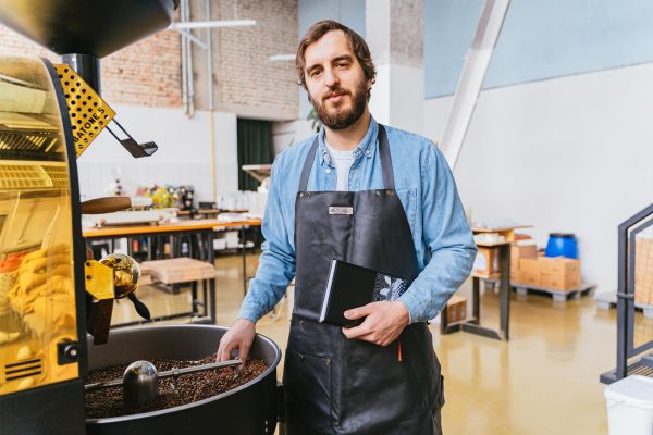 A bearded master roaster stands by a coffee roasting machine, holding a notebook in an industrial workshop.